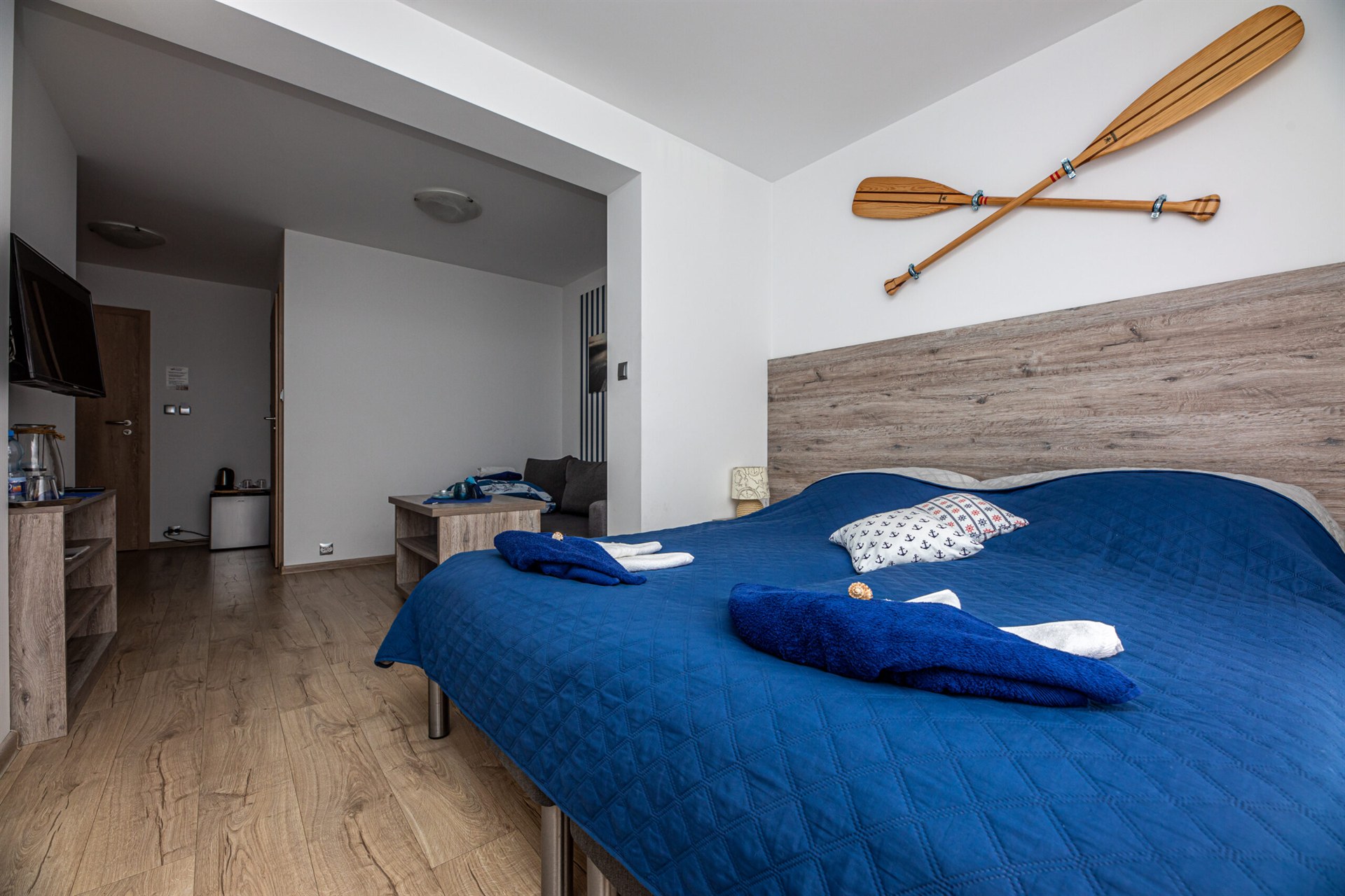 Sypialnia z dużym łóżkiem w granatowym narzucie, złożonymi ręcznikami i skrzyżowanymi drewnianymi wiosłami na ścianie