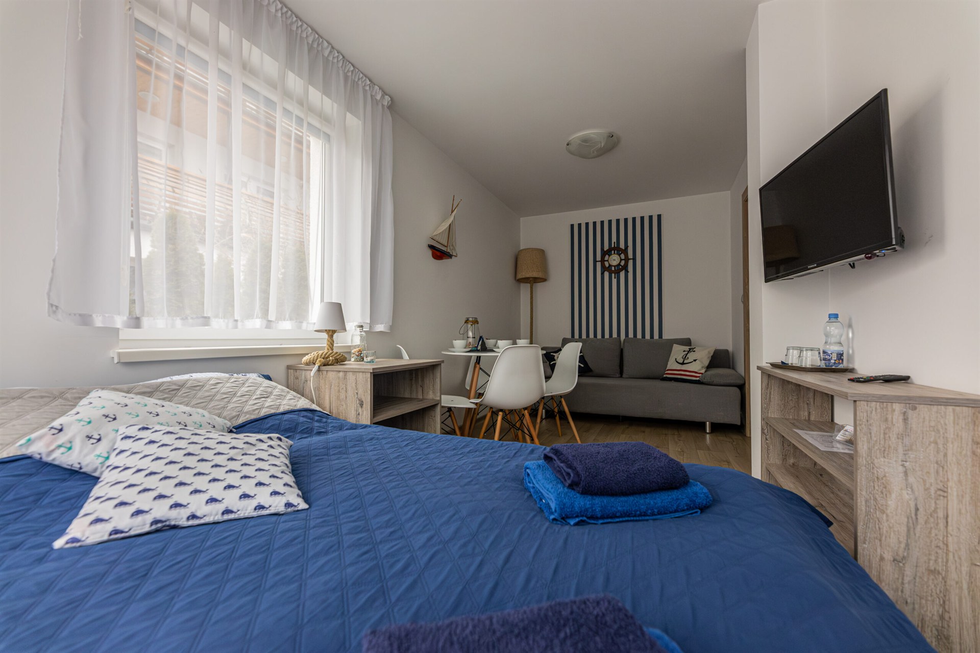 Kawalerka z łóżkiem w granatowej narzucie, szarą sofą, stołem z białymi krzesłami, oknem z białą firaną