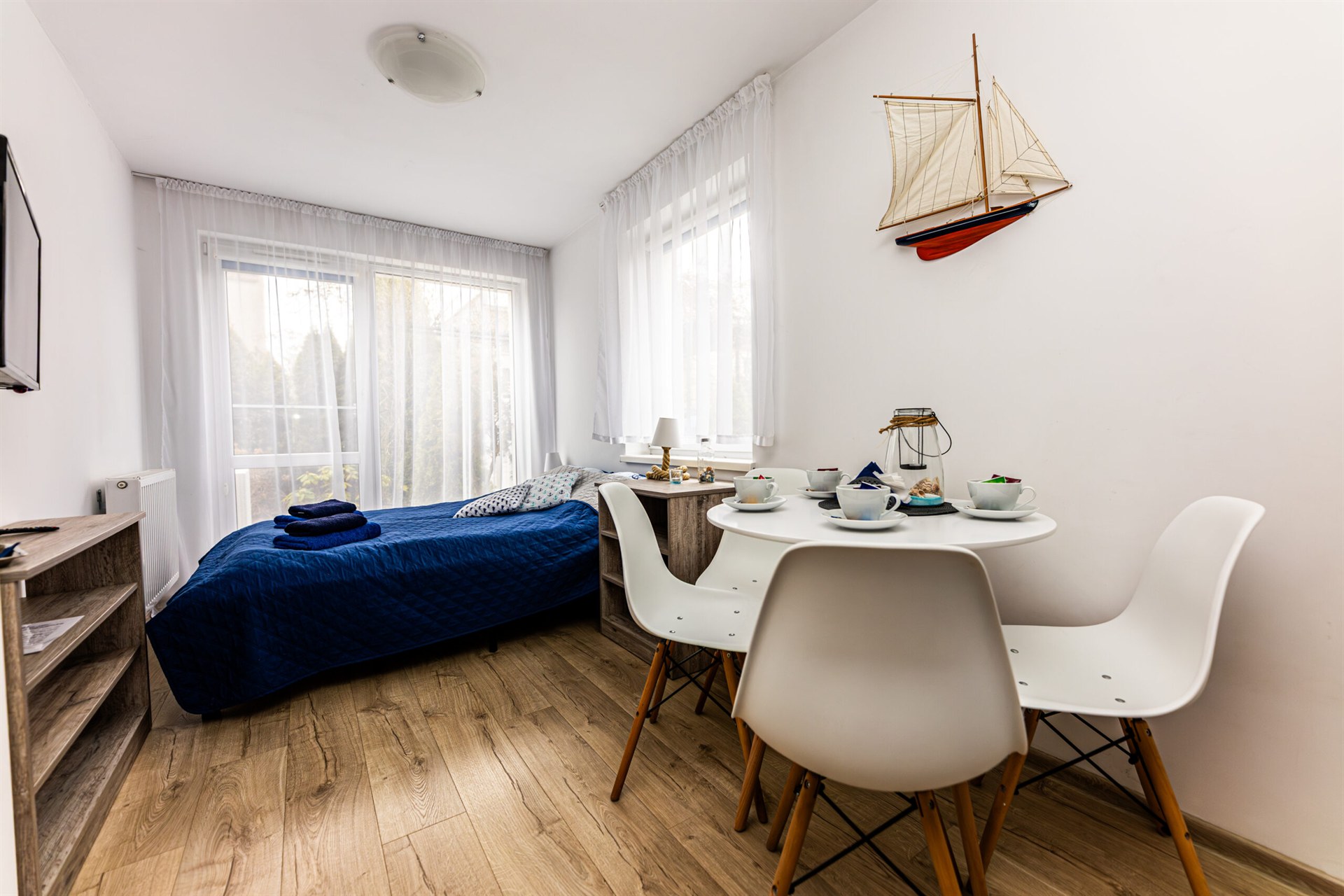 Jasne studio z łóżkiem w granatowej narzucie, okrągłym stołem z białymi krzesłami i zawieszoną makietą żaglowca