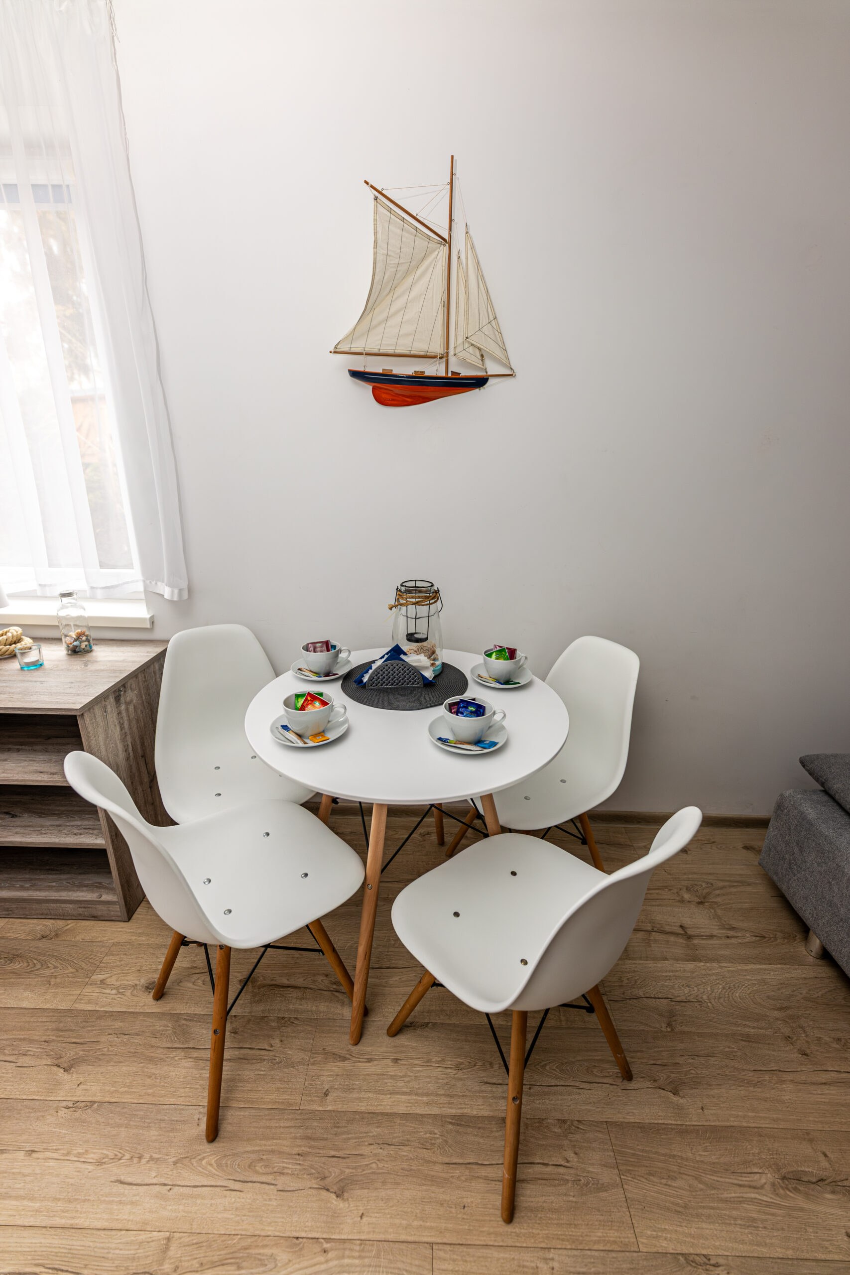 Mały kącik jadalniany: okrągły biały stolik z 4 białymi krzesłami, kolorowe filiżanki i model żaglówki