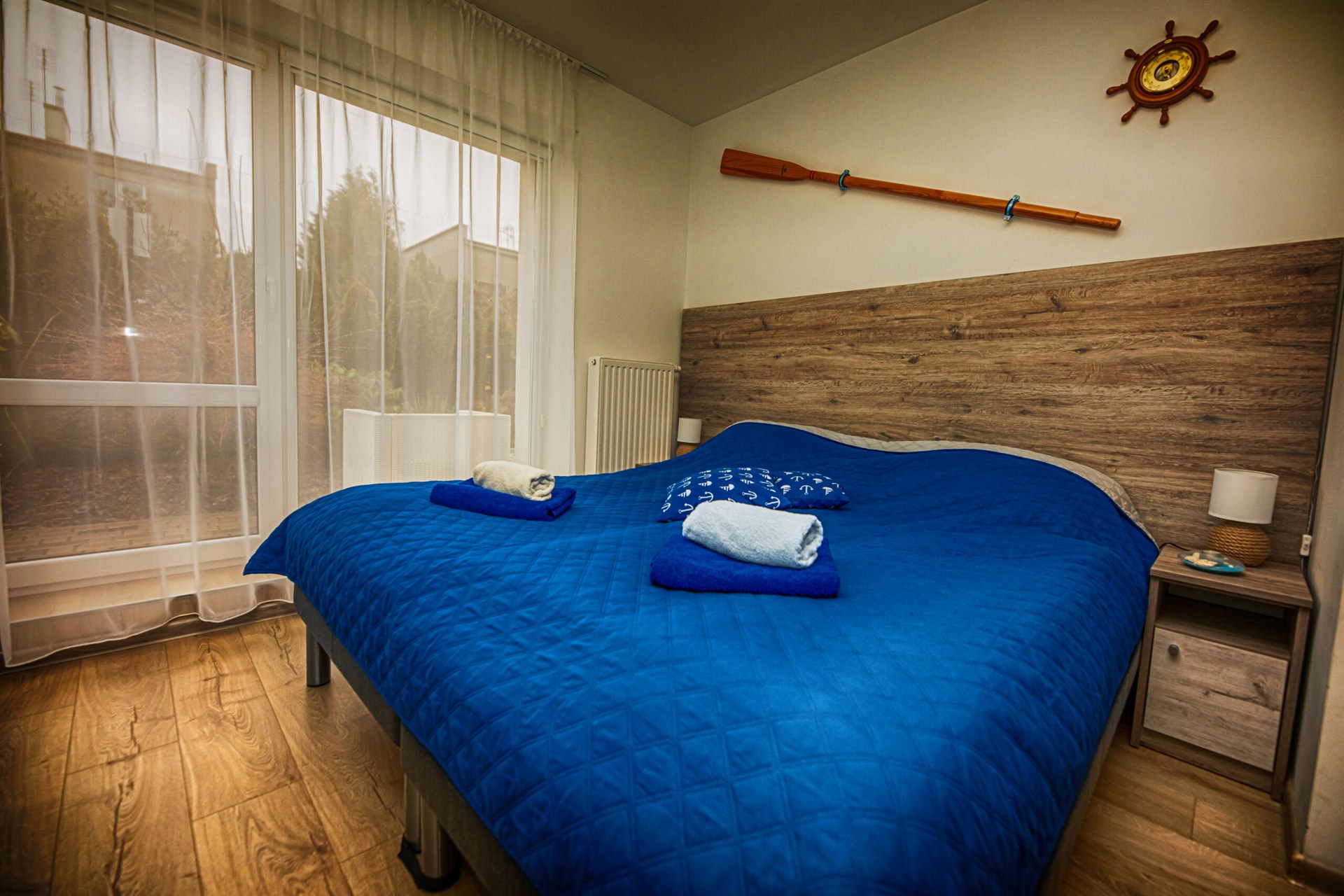 Łóżko z granatową narzutą i ręcznikami, drewniane wezgłowie, wiosło i ster na ścianie, duże okno z firaną