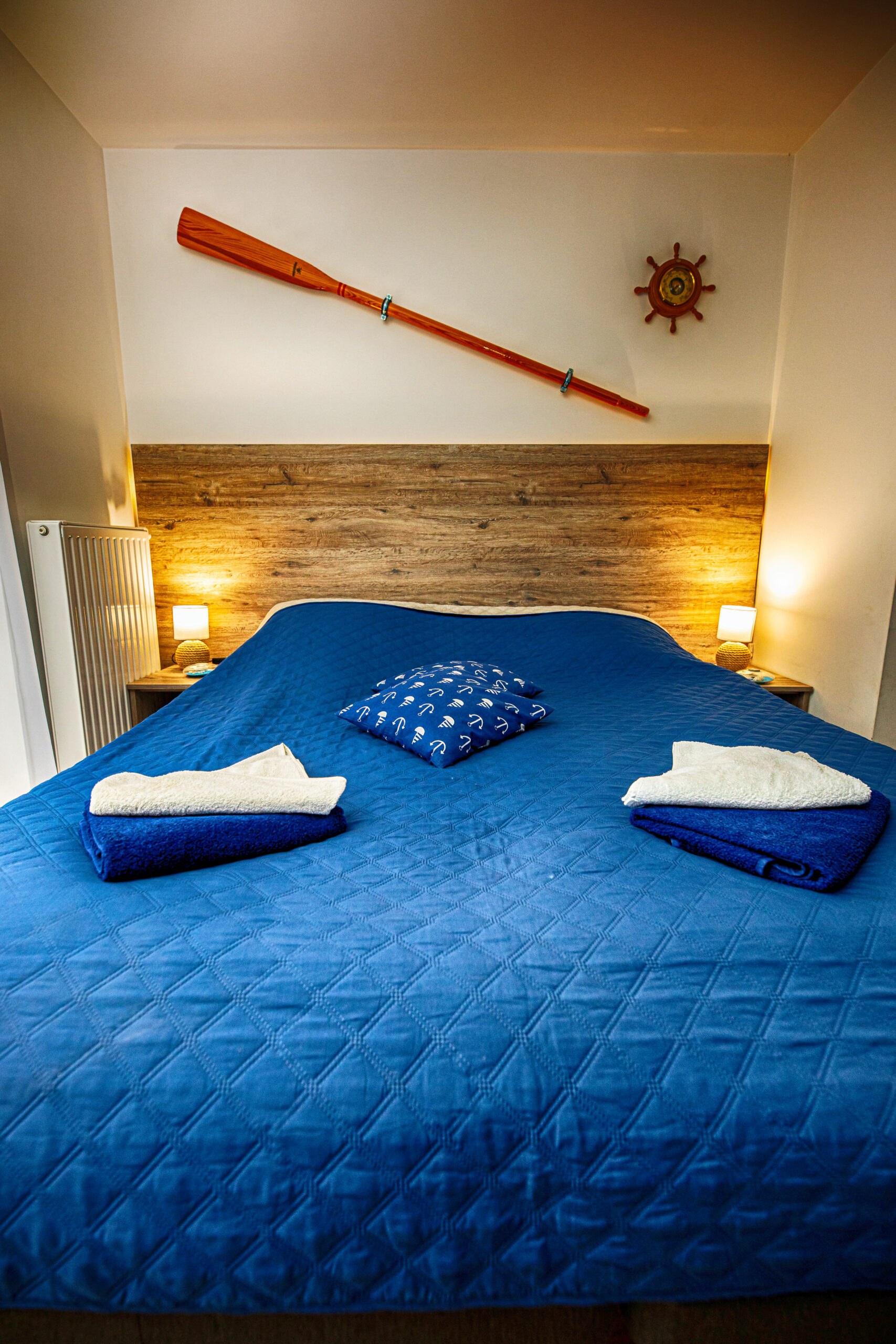 Łóżko z niebieską narzutą, poduszka z kotwicami, złożone ręczniki, drewniane wiosło i koło sterowe na ścianie