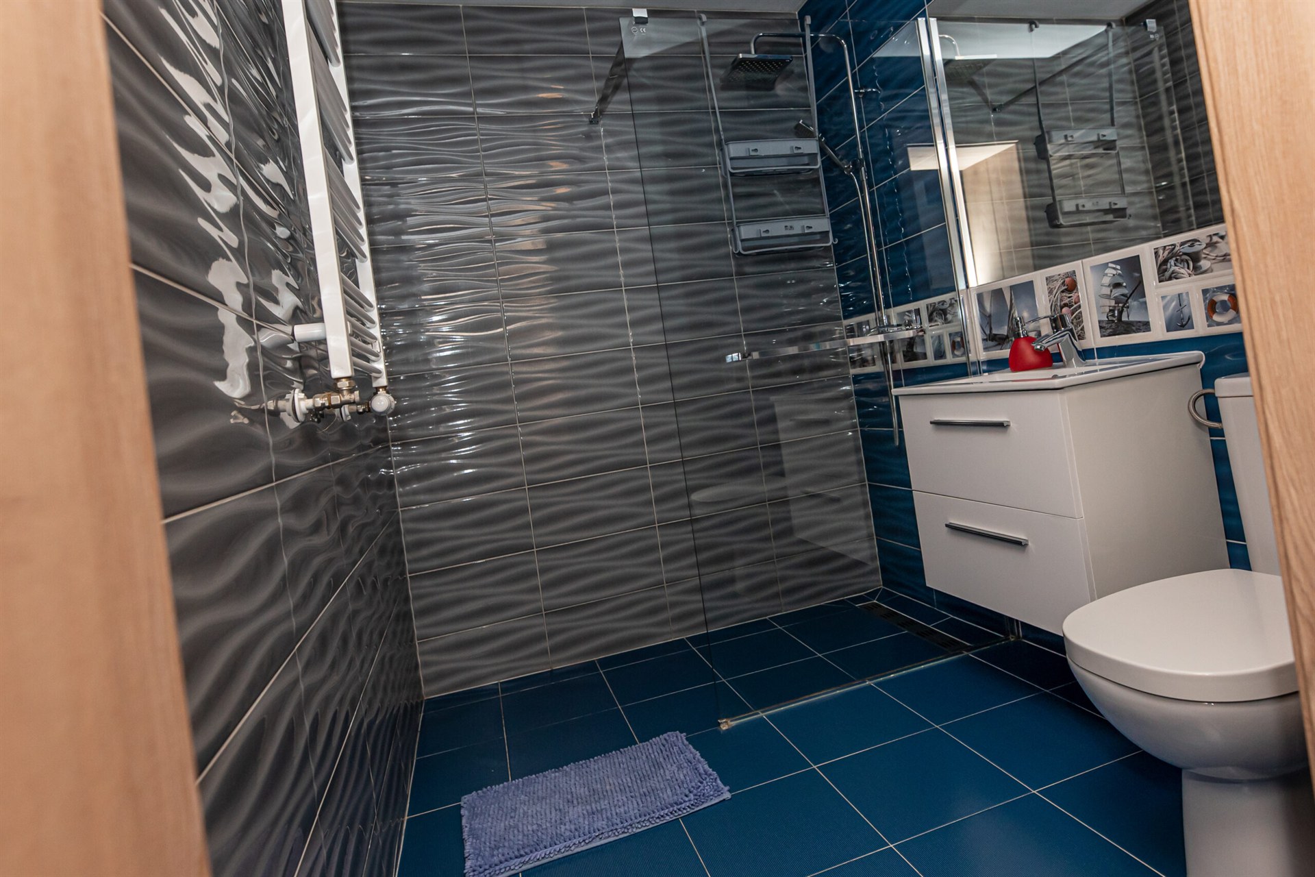 Nowoczesna łazienka z falowanymi szarymi płytkami, niebieską podłogą, przeszklonym prysznicem, białą szafką i czerwonym dozownikiem