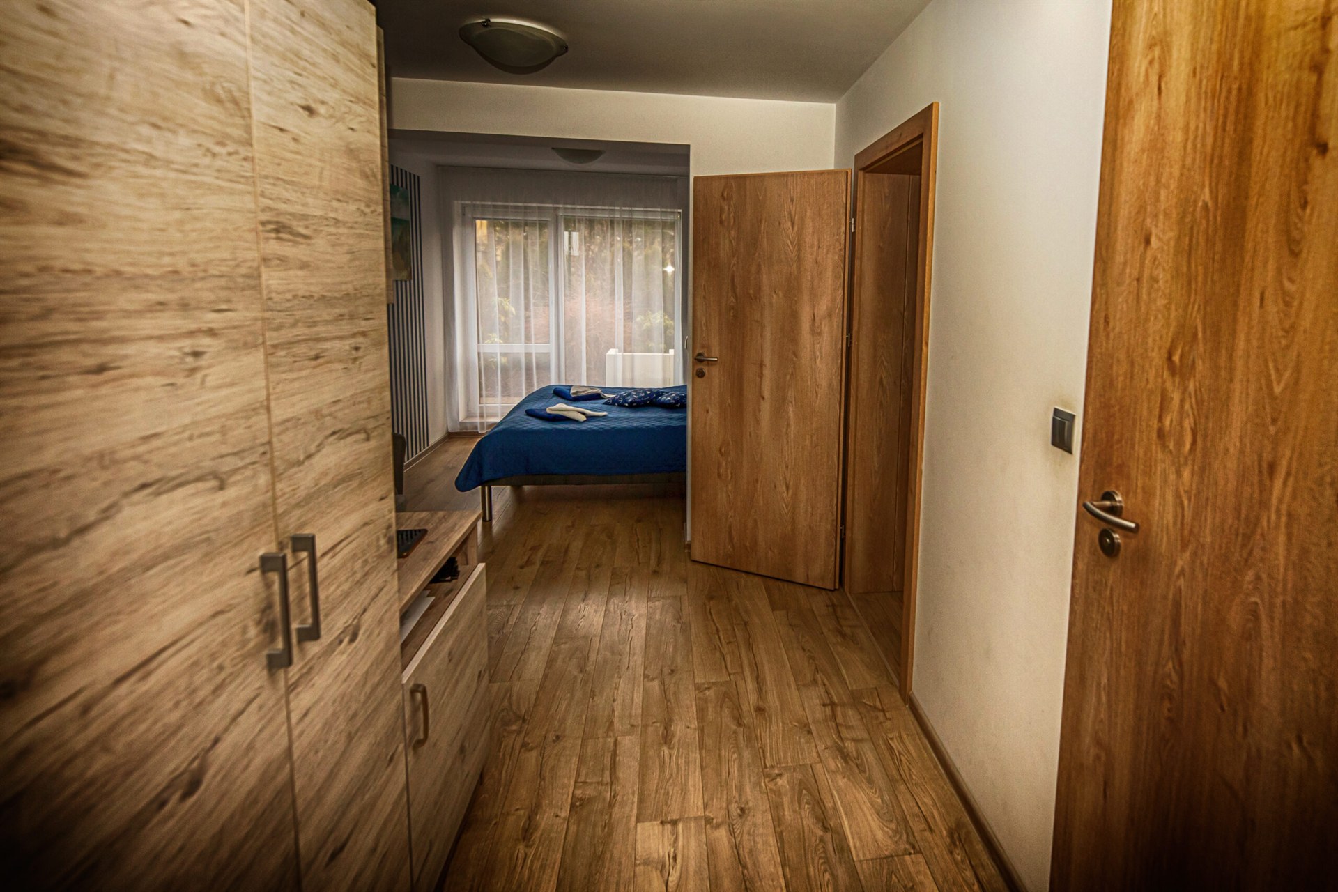 Korytarz z drewnianą podłogą i szafą po lewej prowadzący do sypialni z niebieskim łóżkiem i przezroczystymi zasłonami