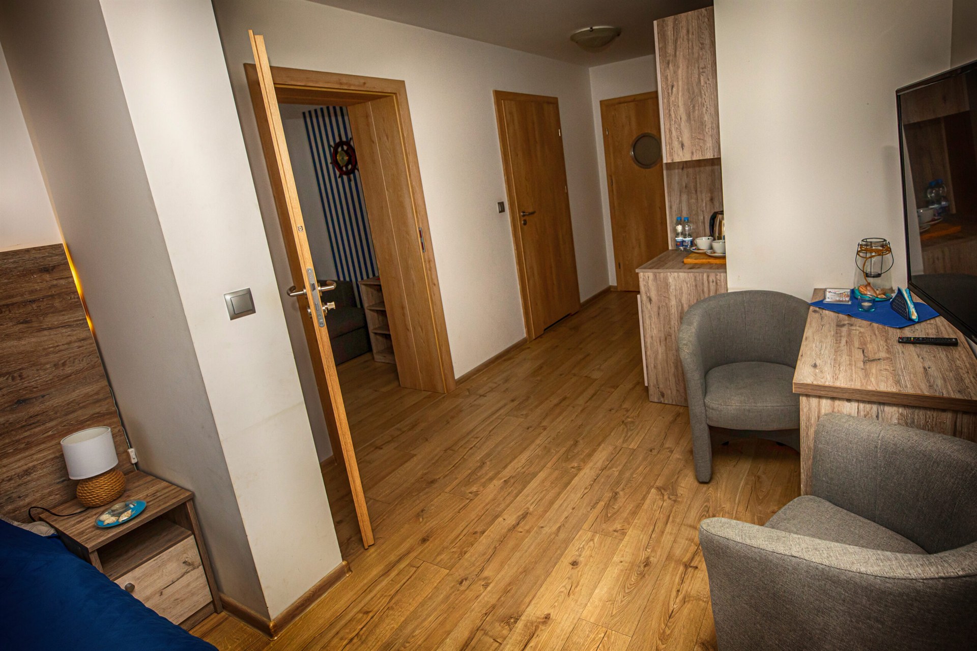 Pokój hotelowy z drewnianą podłogą, biurkiem i telewizorem, dwoma szarymi fotelami i otwartymi drzwiami