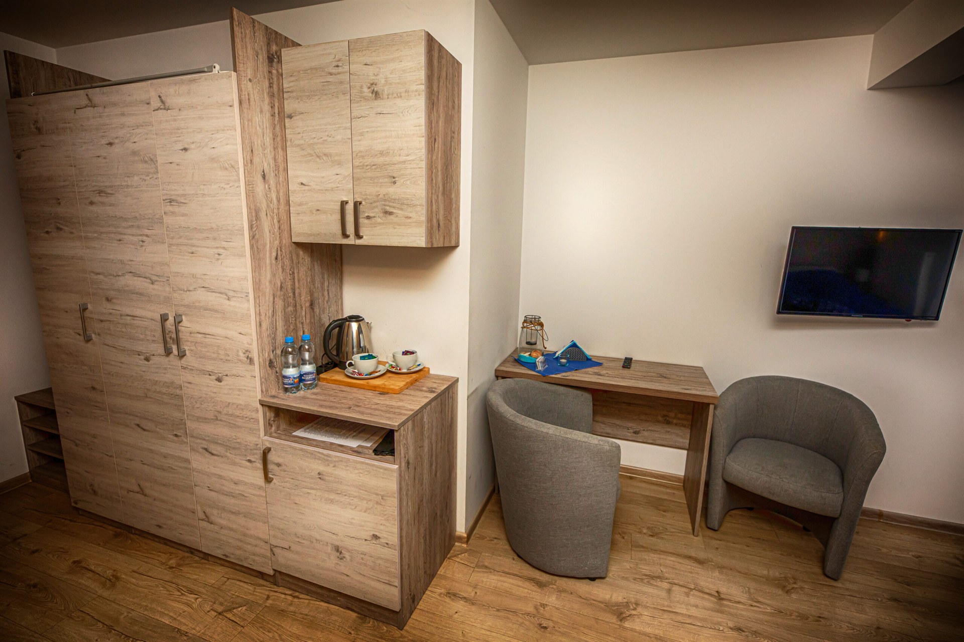 Kąt pokoju z jasną drewnianą szafą i blatem z czajnikiem, biurko z dwoma szarymi fotelami i telewizorem na ścianie