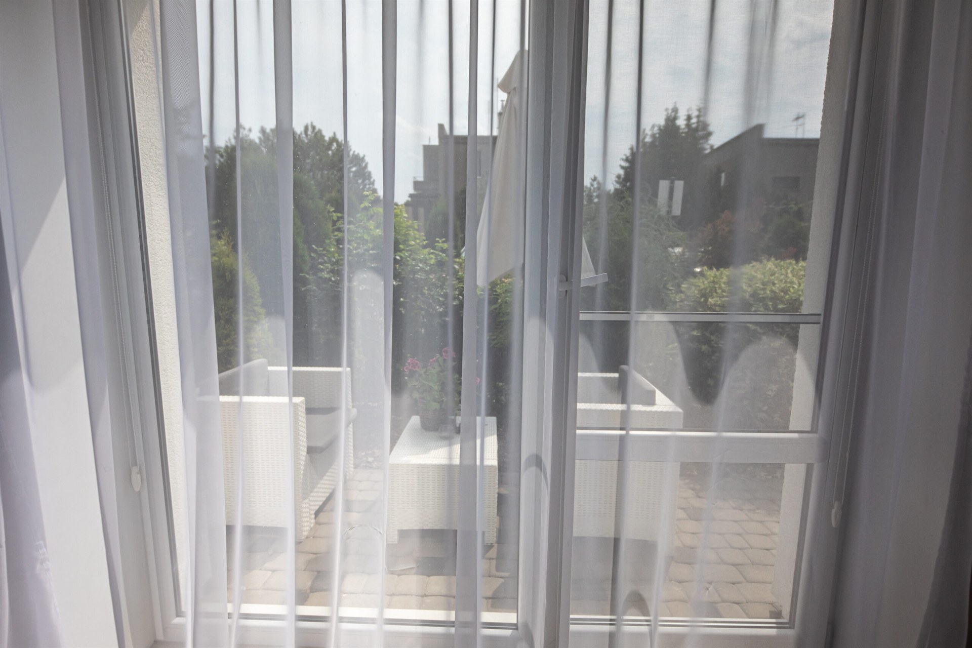 Balkon widziany przez cienką białą firanę, białe rattanowe meble i doniczka z różowymi kwiatami