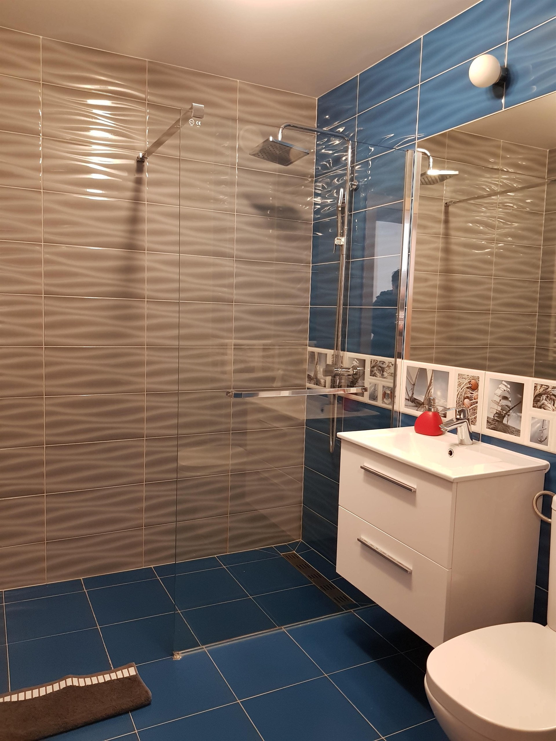 Bezbrodzikowa kabina prysznicowa ze szklaną ścianką, niebieskie płytki i biała szafka z czerwonym dozownikiem