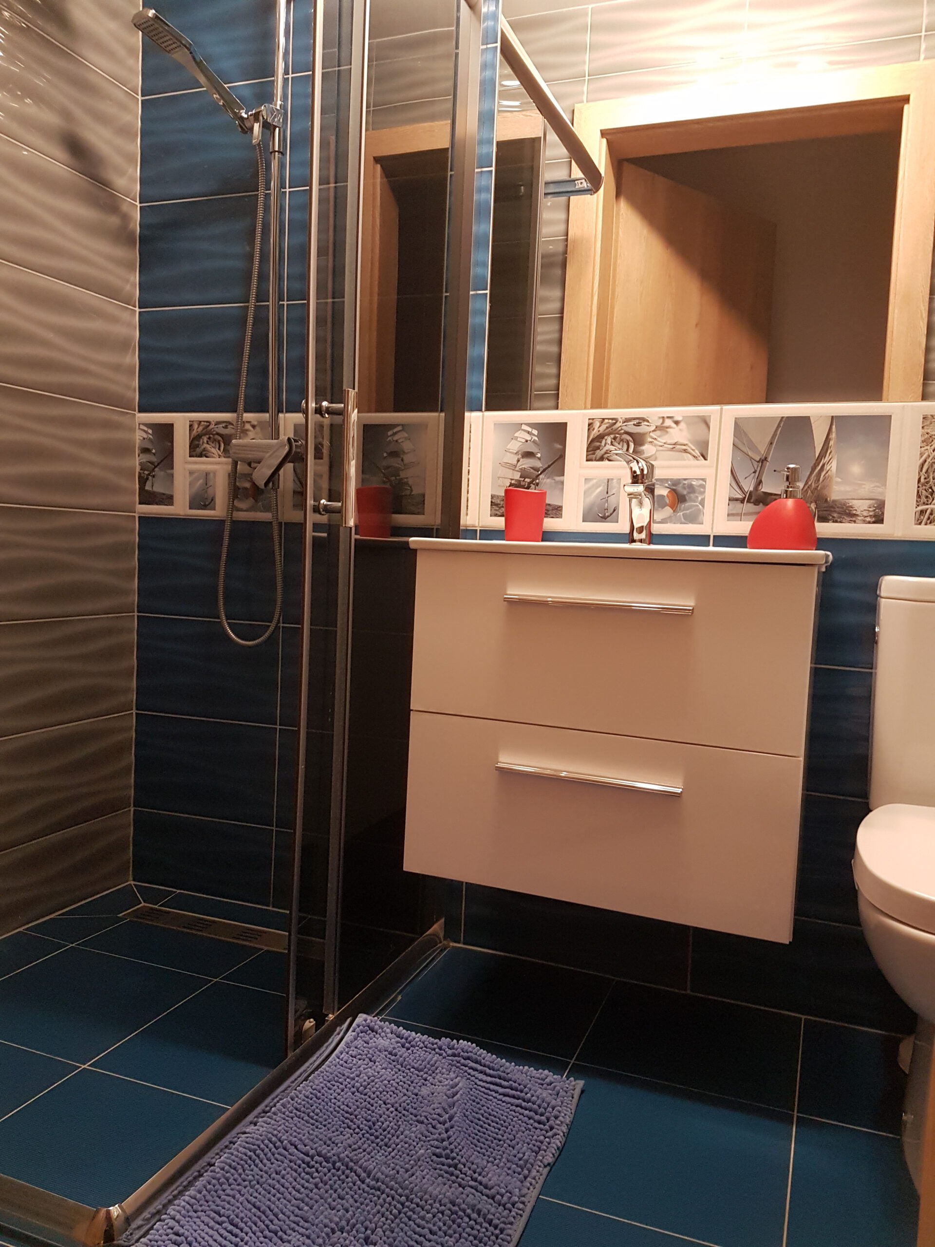 Niebieska łazienka z kabiną prysznicową, białą szafką podumywalkową, czerwonym dozownikiem i niebieską matą