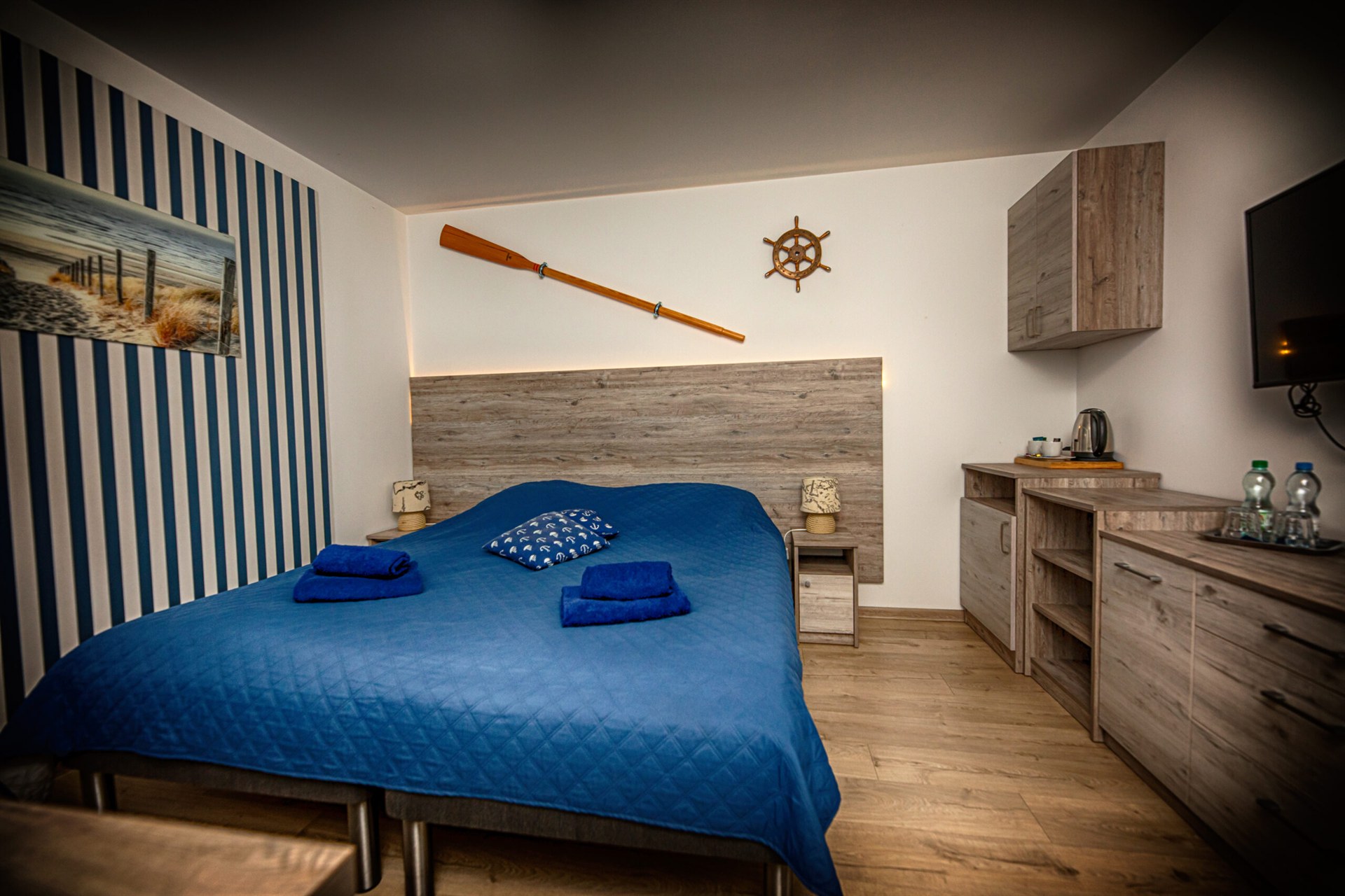 Sypialnia o nadmorskim motywie: duże łóżko z niebieską narzutą, wiosło i ster na ścianie, pasy i drewniane meble