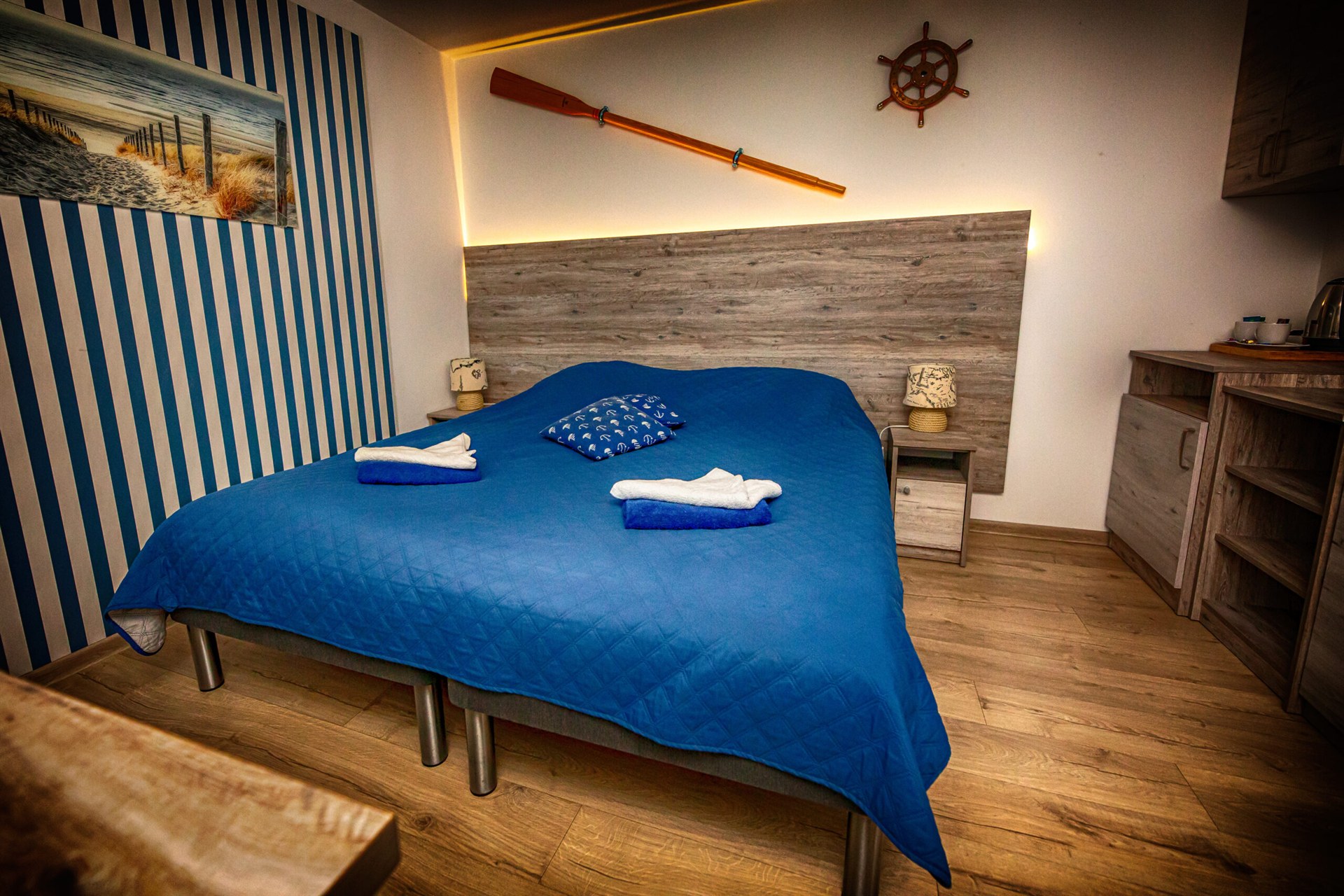 Sypialnia marynistyczna z niebieską narzutą, pasiastą ścianą, wiosłem i sterem oraz złożonymi ręcznikami