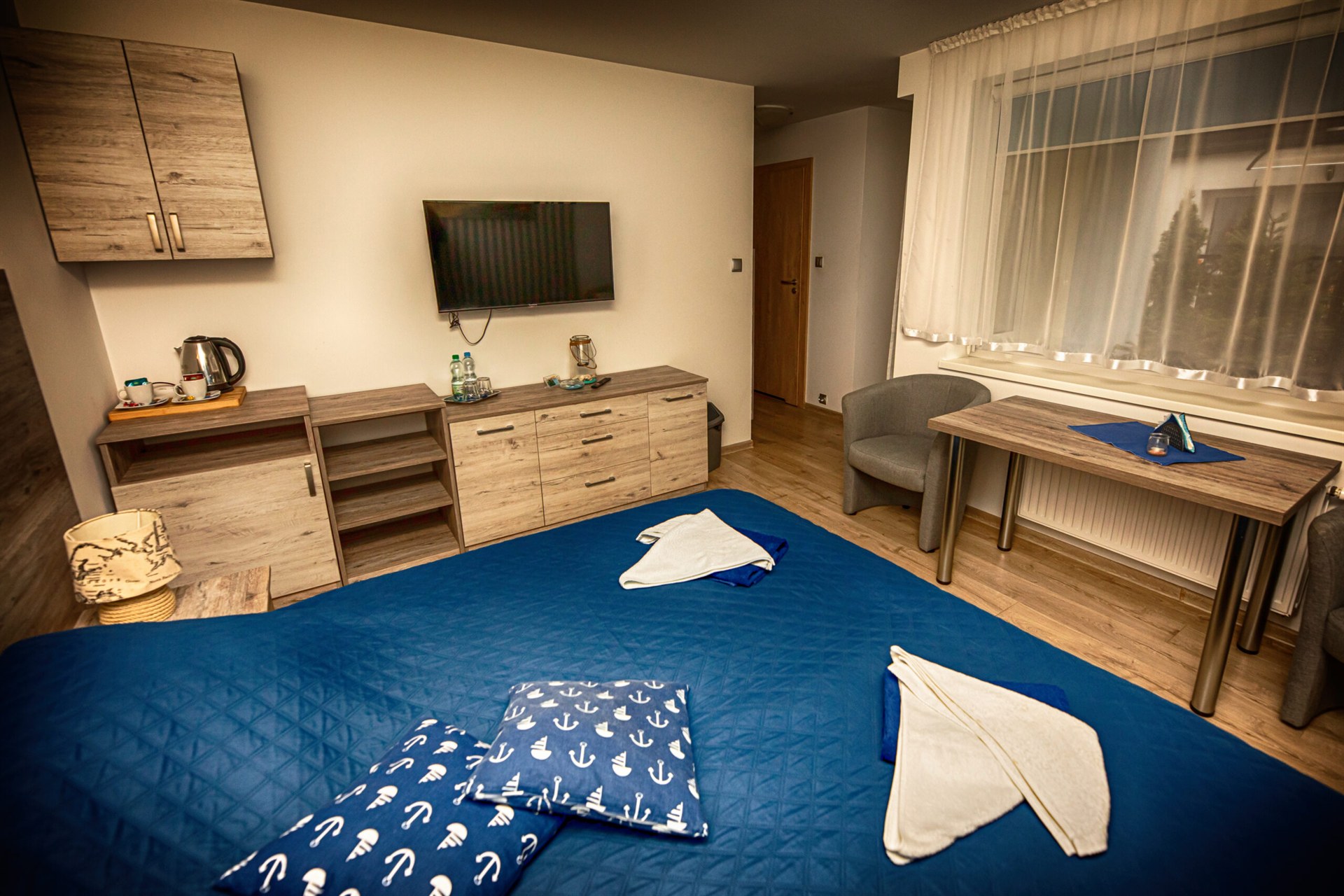 Pokój z niebieskim łóżkiem, poduszkami w kotwice, drewnianymi szafkami, telewizorem na ścianie i stolikiem przy oknie