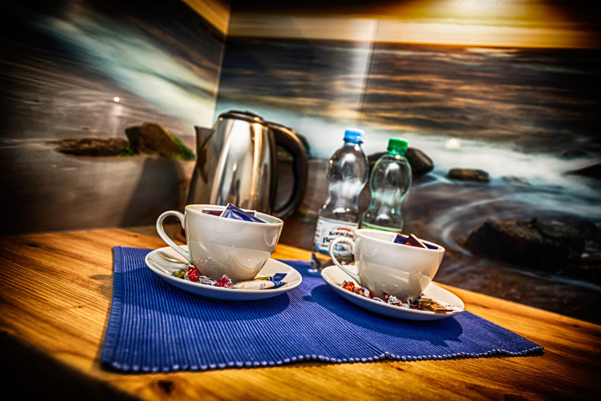 Dwie białe filiżanki z cukierkami na talerzykach na niebieskiej podkładce obok czajnika i butelek