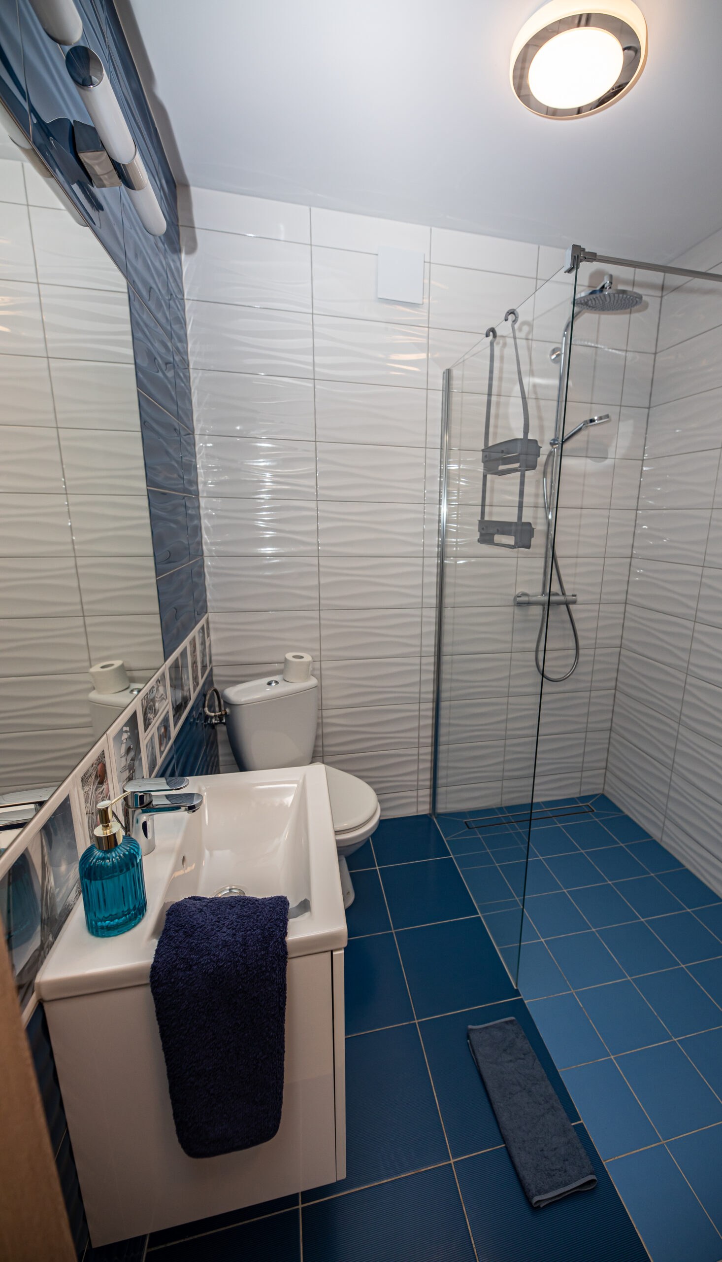 Łazienka z falistymi białymi płytkami, niebieską podłogą, szklanym prysznicem i umywalką z turkusowym dozownikiem