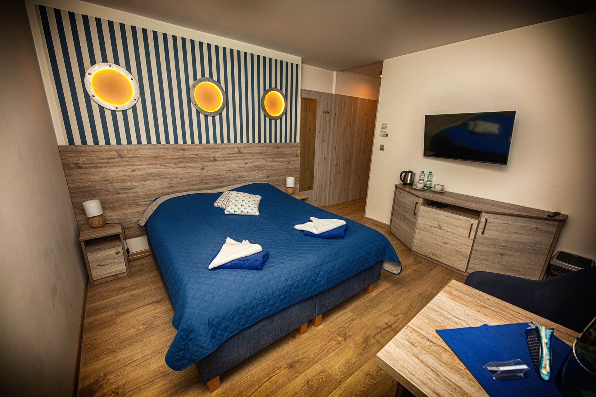 Sypialnia z dużym łóżkiem w granatowej narzucie, trzema okrągłymi podświetlanymi okienkami na pasiastym tle