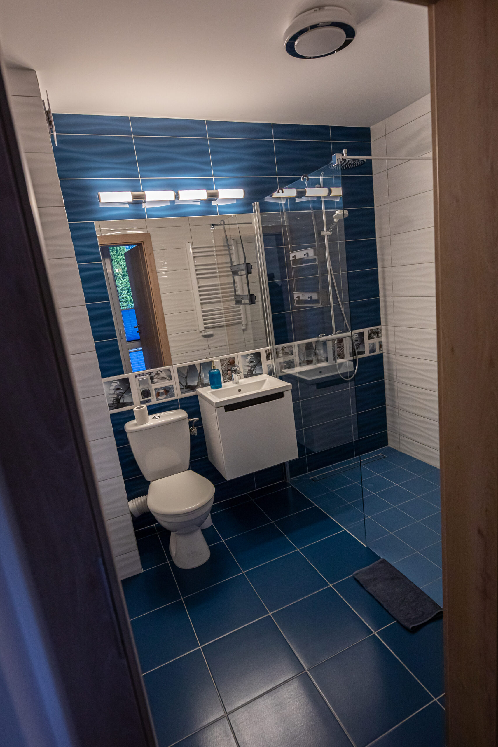 Łazienka z granatowymi płytkami, białą toaletą, podwieszaną umywalką i przeszkloną kabiną prysznicową