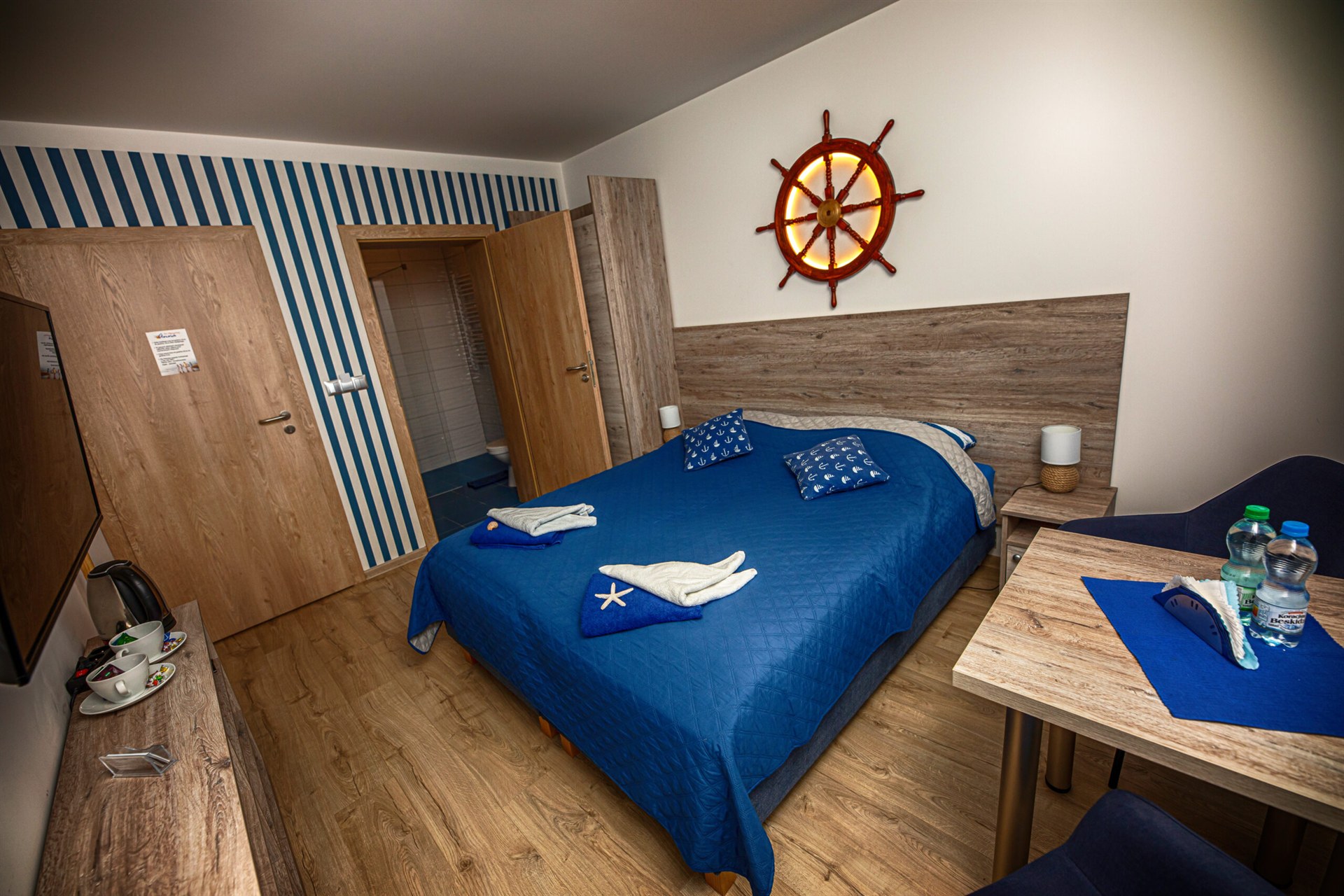 Sypialnia z granatową pościelą, drewnianym wezgłowiem, podświetlanym kołem sterowym i niebiesko-białą pasiastą ścianą