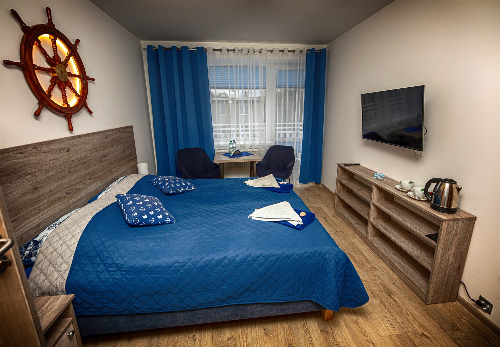 Pokój hotelowy z dużym łóżkiem w niebieskiej narzucie, sterem okrętowym na ścianie, dwoma fotelami przy oknie i telewizorem