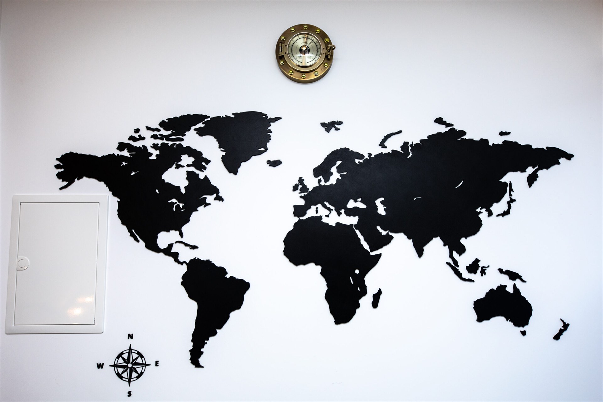 Czarna mapa świata na białej ścianie z miedzianym zegarem portowym u góry i kompasem u dołu po lewej