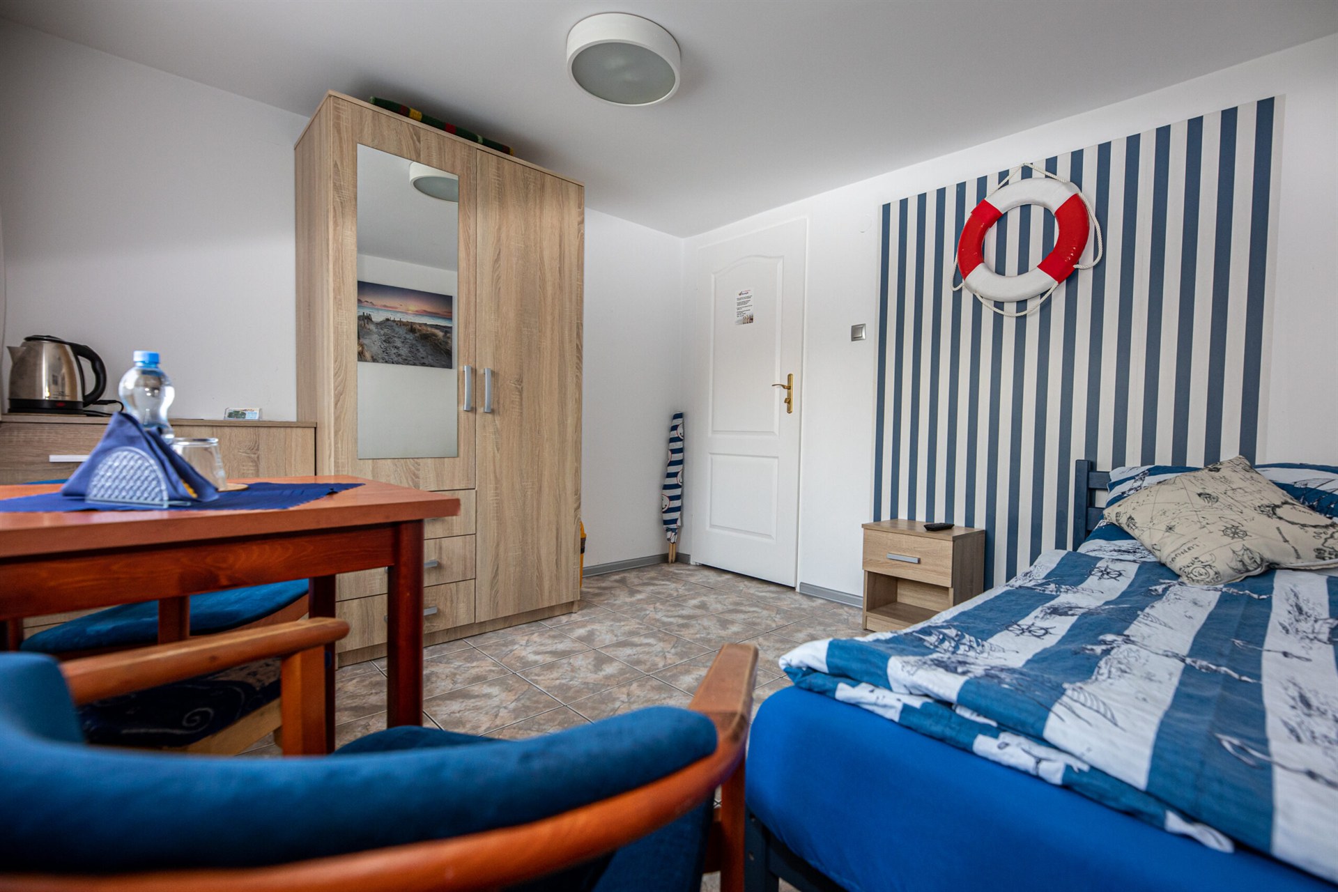 Niewielki pokój morski: łóżko z granatowo-białą pościelą, pasiasta ściana i czerwono-białe koło ratunkowe