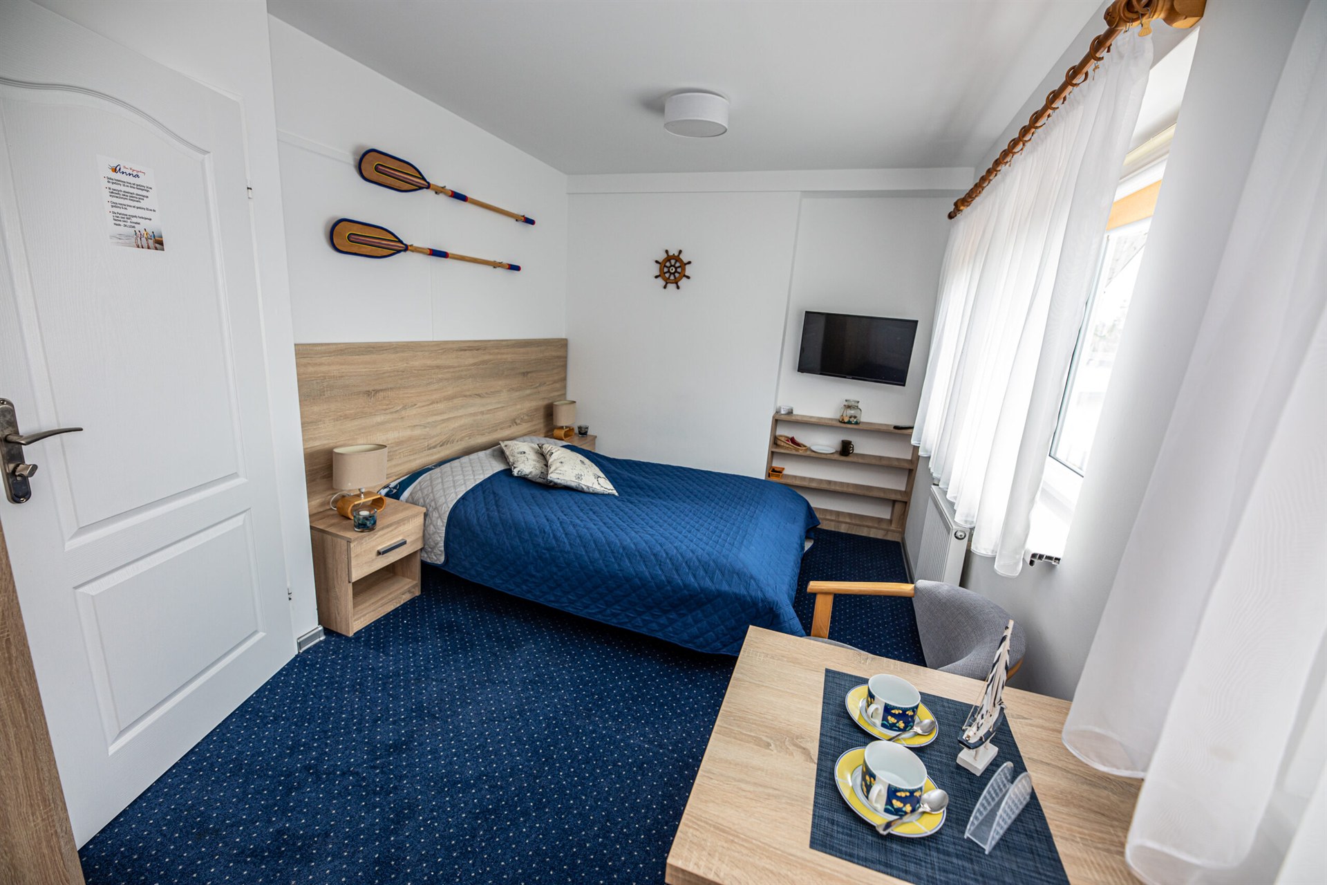 Sypialnia marynistyczna: granatowa pościel, drewniane wiosła i koło sterowe, stolik z filiżankami