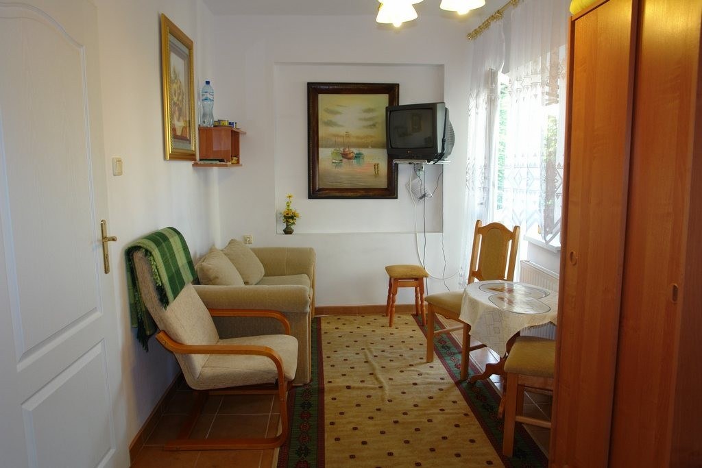 Mały salon z beżowymi fotelami i kanapą, zielonym kocem, wiszącym telewizorem CRT, stołem przy oknie i drewnianą szafą