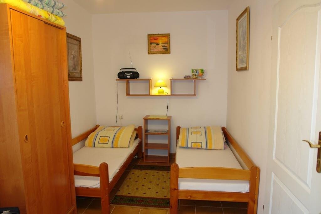 Dwa drewniane łóżka z żółto-niebieskimi poduszkami, między nimi półka z lampką i radiem