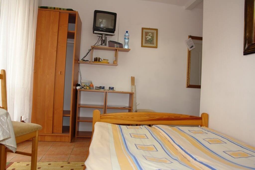 Mała sypialnia z drewnianym łóżkiem w paskowanej żółto-niebieskiej pościeli, szafa, półki i stary telewizor na ścianie