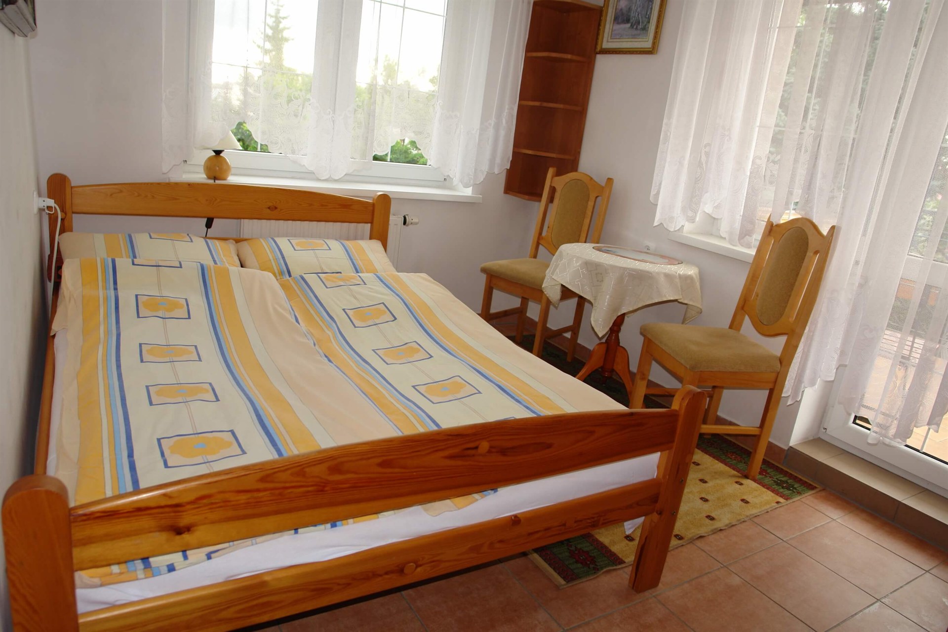 Drewniane łóżko z żółto-niebieską pościelą przy oknie z koronkowymi firankami i stolikiem z dwoma krzesłami