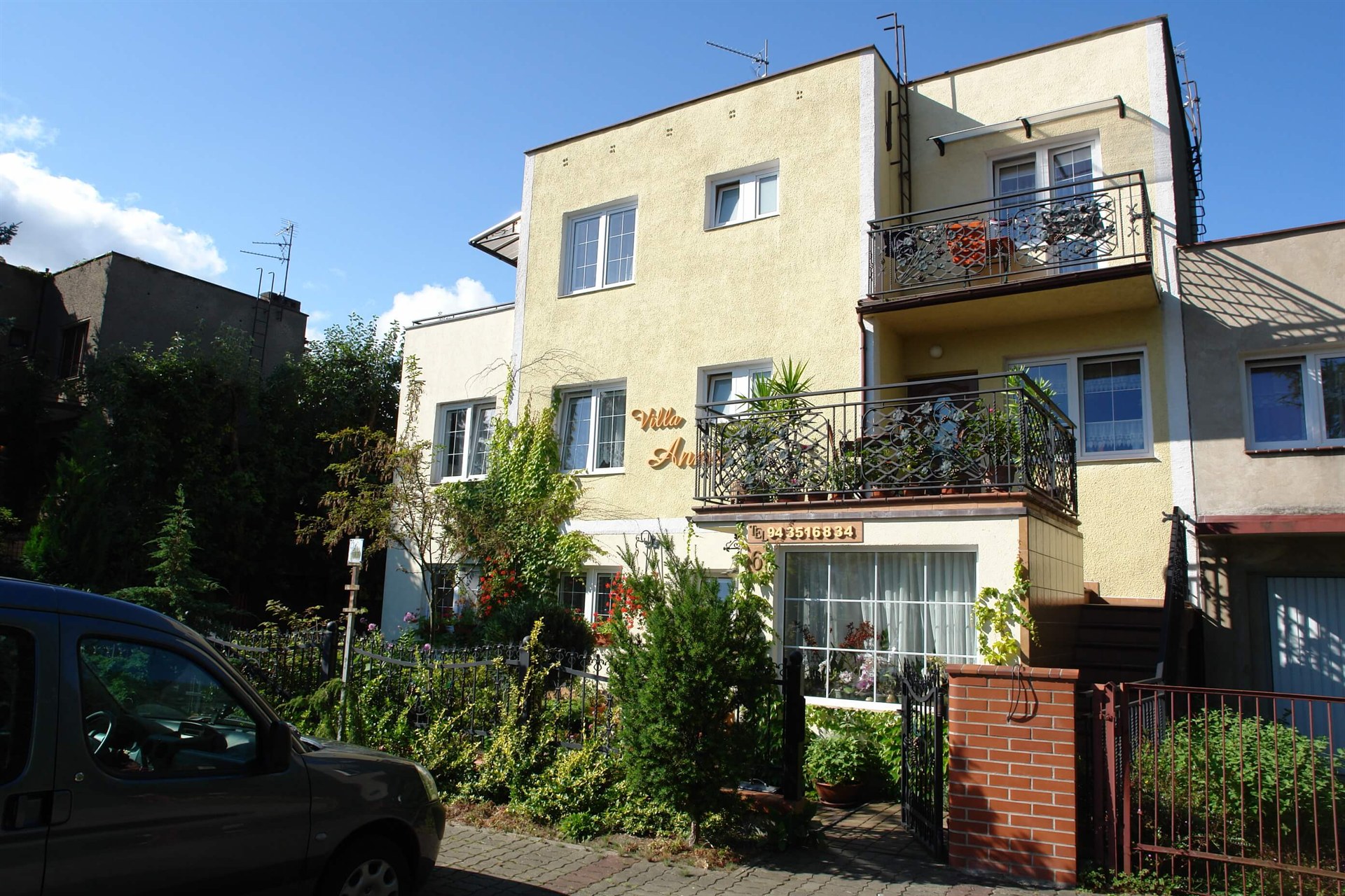 Żółta willa z ozdobnymi balkonami, bujnym ogrodem przy furtce i zaparkowanym samochodem